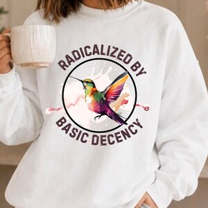 May include: White sweatshirt with a vibrant hummingbird illustration inside a black circle. The text "RADICALIZED BY BASIC DECENCY" is arranged around the circle. The hummingbird is depicted with green, yellow, purple, and pink feathers.