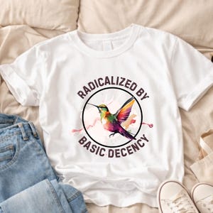 May include: White t-shirt with a vibrant hummingbird illustration inside a black circle, accompanied by the text "RADICALIZED BY BASIC DECENCY". The hummingbird displays green, yellow, purple, and orange colours. The shirt is shown with blue jeans and white trainers.