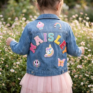 Peut inclure: Une veste en jean bleu ornée de patchs colorés au dos, incluant un cupcake, un arc-en-ciel, une licorne, un visage souriant et un papillon. Le nom MAISLEY est écrit en grandes lettres en tissu pailleté multicolore au centre du dos.