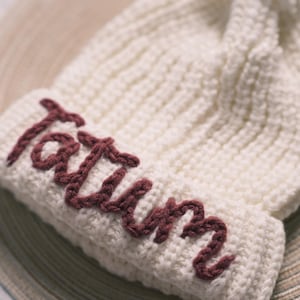 Può includere: Berretto lavorato a maglia color crema con il nome "Tatum" ricamato in bordeaux. Il cappello è appoggiato su una superficie chiara e strutturata. Il berretto ha un design a coste.