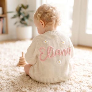 May include: A cream-colored knit sweater featuring the name Eliana embroidered in pink yarn across the back. The sweater is decorated with small, white, daisy-like floral accents with yellow centers scattered around the pink lettering.