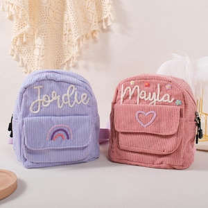 May include: Two small corduroy backpacks, one lavender and one pink. The lavender backpack has the name "Jordie" embroidered on it, along with a rainbow. The pink backpack has the name "Mayla" embroidered, with a heart and flowers.