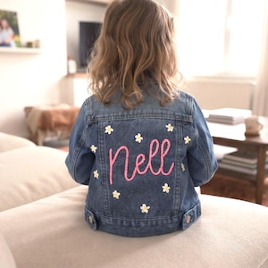 May include: A blue denim jacket featuring the name Nell embroidered in pink script on the back. The jacket is decorated with small white and yellow flower embroidery scattered across the back panel, including long sleeves and a buttoned hem.