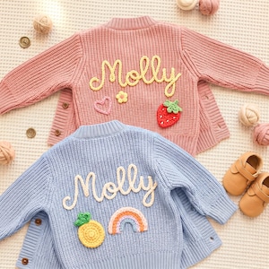 May include: Two knitted children's cardigans, one pink and one blue, with the name "Molly" in yellow script. The pink cardigan has a strawberry, heart, and flower applique. The blue cardigan has a pineapple and rainbow applique. Both have button fastenings.