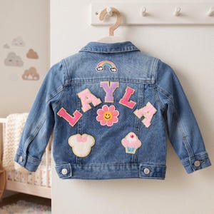 May include: A blue denim jacket featuring colourful felt patches on the back. The name LAYLA is spelled out in large pink and purple letters, accompanied by a rainbow, a smiling pink flower, a white mouse-shaped silhouette, and a cupcake patch.