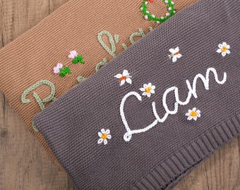 Puede incluir: Se muestran dos mantas de punto, una beige y otra gris. La manta beige tiene el nombre "Richard" bordado en verde con detalles de flores rosas. La manta gris presenta el nombre "Liam" en blanco con detalles de margaritas.