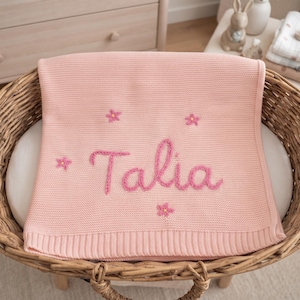 Könnte beinhalten: Eine hellrosa gestrickte Babydecke mit dem in dunkelrosa Garn eingestickten Namen Talia in der Mitte. Die Decke ist mit kleinen, verstreuten rosa Blumenmotiven mit gelben Zentren verziert und verfügt über einen gerippten Randabschluss.