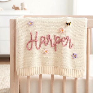 Op de afbeelding: Een crèmekleurige gebreide deken met de naam Harper geborduurd in roze garen. De deken is versierd met kleine, driedimensionale gehaakte bloemen in roze en paarse tinten, samen met een kleine, zwart-gele hommel.