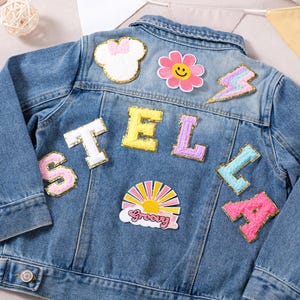 May include: A light blue denim jacket adorned with various colourful patches. Patches include a white bow, a pink flower with a smiling face, a lightning bolt, and the name "STELLA" in individual letters. The jacket also features a "Groovy" patch.