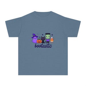 May include: A slate blue short-sleeve t-shirt featuring a graphic of a purple ghost in a witch hat, an orange pumpkin, a black cat, a green monster, and a candy corn character with the text "bootastic" printed in dark purple below them.