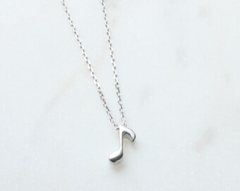 May include: A silver-colored necklace featuring a small musical eighth note pendant. The pendant hangs from a delicate chain, creating a minimalist and elegant design. The necklace is displayed on a white marble surface.
