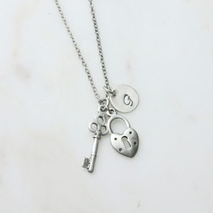 Może przedstawiać: Srebrny naszyjnik z sercowatym zamkiem, kluczem i okrągłym wisiorkiem z wygrawerowaną literą "S".