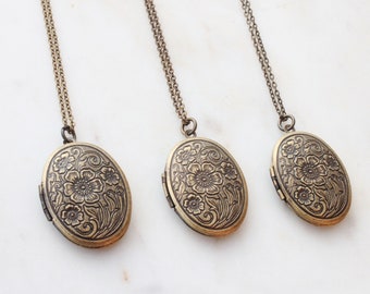 May include: Three antique bronze oval lockets with floral designs hang from delicate chains. The lockets are closed, and the intricate floral patterns are visible on the front. The background is a white surface.