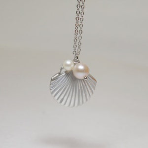 Puede incluir: Un collar de plata con un colgante de concha y tres perlas blancas. La concha tiene una superficie texturizada y está detallada con líneas.