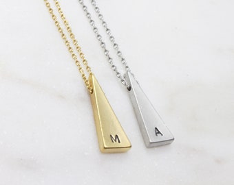 May include: Two pendant necklaces with a triangular design. One is gold-toned with the letter "M", the other is silver-toned with the letter "A". Both hang from delicate chain necklaces. The background is a white marble surface.