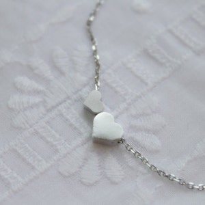 May include: A silver necklace with two heart-shaped charms. The necklace is on a white background.