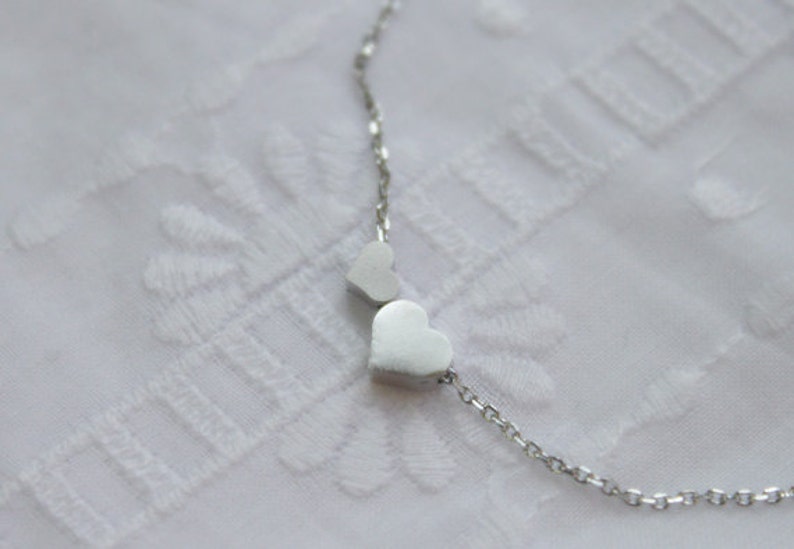May include: A silver necklace with two heart-shaped charms. The necklace is on a white background.