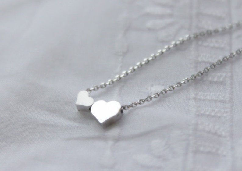 May include: A silver necklace with two small heart charms. The necklace is on a white fabric background.