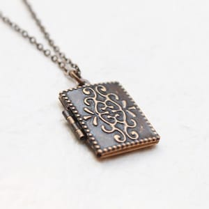 Könnte beinhalten: Eine silberne Kettenkette mit einem kleinen, rechteckigen Anhänger. Der Anhänger ist aus dunklem Metall mit einem dekorativen, goldfarbenen Design.