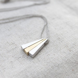 Könnte beinhalten: Eine silberne Kettenkette mit einem dreieckigen Anhänger aus drei übereinanderliegenden Metallstäben. Der obere Stab ist golden, der mittlere Stab ist silber und der untere Stab ist silber.
