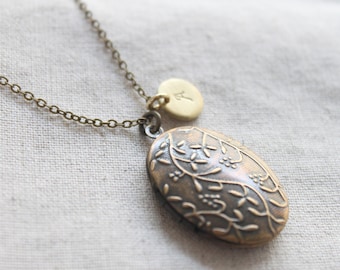 May include: A gold necklace with a delicate chain and a small oval locket. The locket has a floral design and is attached to a round charm with the letter "A" engraved on it.