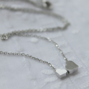 May include: A silver chain necklace with two silver heart charms. The larger heart charm is slightly offset from the smaller heart charm.