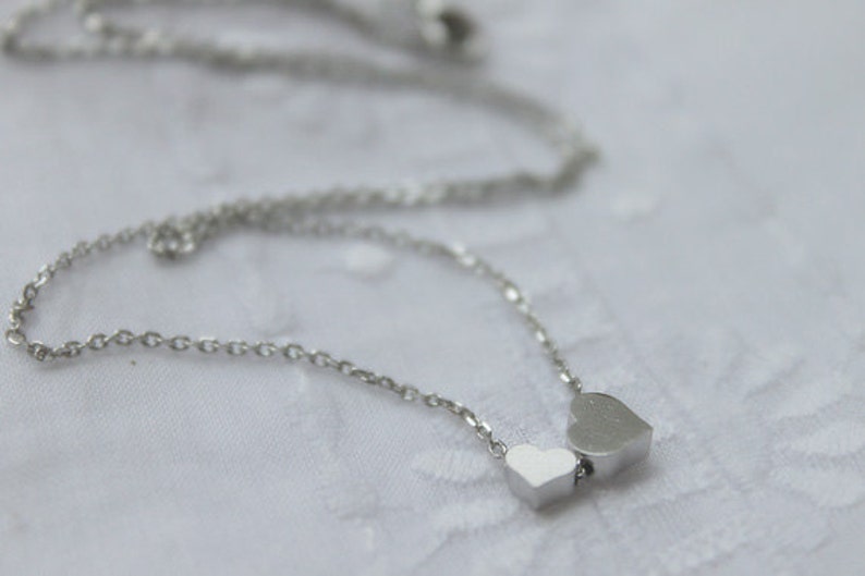May include: A silver chain necklace with two silver heart charms. The larger heart charm is slightly offset from the smaller heart charm.