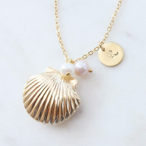 Puede incluir: Collar de oro con un colgante de concha, detalles de perlas y un pequeño dije de disco de oro con la letra "R" grabada.