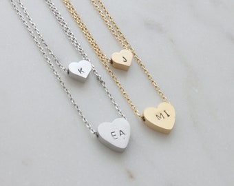 May include: Three silver and gold necklaces with heart-shaped pendants. The pendants are engraved with the letters "K", "J", and "MI".