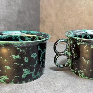 May include: Two ceramic mugs with a unique handle design. The mugs are black with a green speckled pattern and a green rim. The handles feature two finger holes. The mugs are suitable for coffee, tea, or other beverages.