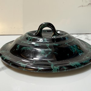 May include: A black ceramic lid with a green marbleized pattern and a rounded handle. The lid is circular and slightly domed, with a glossy finish. The handle is a loop shape, and the overall design is elegant.