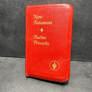 May include: A small, red book with gold lettering. The cover reads "New Testament Psalms Proverbs." A gold emblem of a chalice is at the bottom. The book is standing upright.