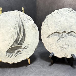 May include: Two decorative, round, off-white stone plaques. One features a sailboat carving, the other a seagull in flight. Each plaque is displayed on a stand, one gold-toned, the other black.
