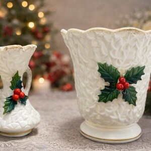 May include: Two white ceramic vases with a textured, embossed leaf pattern and scalloped rims. The smaller vase features two handles, while both pieces are decorated with a central holly motif consisting of green leaves and three bright red berries.