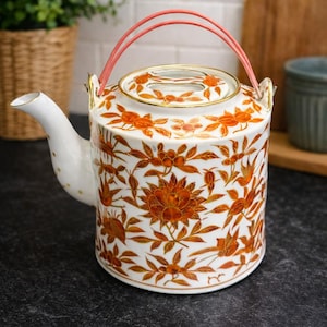 May include: White ceramic teapot with a floral pattern in orange and gold. The teapot has a pink handle and a gold rim. The spout is white with gold polka dots. A decorative kitchenware item.