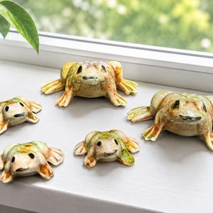 May include: Five ceramic frog figurines in varying sizes are arranged on a white windowsill. The frogs have a tan and green color scheme with detailed features. A potted plant is visible in the background.