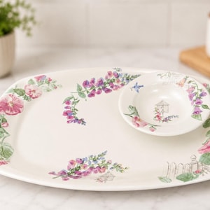May include: A white ceramic serving platter and a matching small bowl featuring a watercolor-style floral pattern with pink and purple flowers, green leaves, a white picket fence, and a small birdhouse illustration on the surface of both pieces.