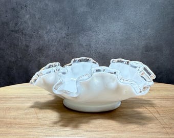 May include: A white, ruffled-edge glass bowl with a pedestal base. The bowl has a wavy, scalloped rim with a clear, frosted edge. The bowl is sitting on a wooden surface.
