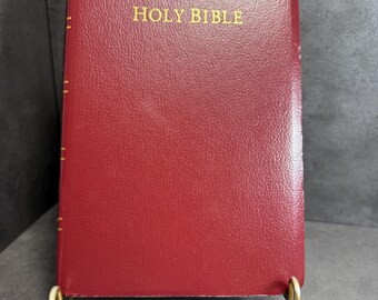 May include: A burgundy Holy Bible with a textured, leather-like cover and gold-colored lettering on the front. The spine features gold-colored horizontal lines. The book is displayed on a gold-colored metal stand with a small white tag labeled #9.