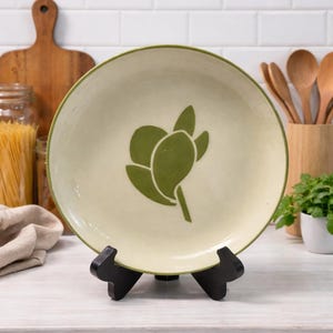 May include: A ceramic plate with a light cream background and a green floral design. The plate has a green rim and is displayed on a black stand. Kitchenware.