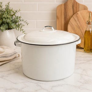 May include: A white enamel cooking pot with a lid, featuring a black trim and handles. The pot is on a marble countertop, with a white tile background. Kitchenware.