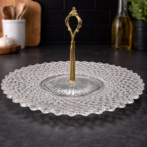 May include: A clear, two-tiered glass serving tray with a gold-colored handle. The tray has a textured, patterned surface and a scalloped edge. Ideal for displaying desserts or appetizers.