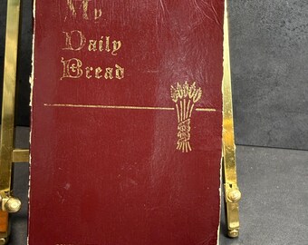 May include: A burgundy-colored book with gold lettering and a gold illustration of wheat. The text on the cover reads "My Daily Bread" and "CONFRATERNITY OF THE PRECIOUS BLOOD". The book is displayed on a gold easel.