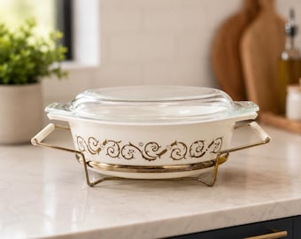 May include: A vintage white casserole dish with a clear glass lid, featuring a gold floral design. It sits in a gold-toned metal stand with white handles. The dish is on a white countertop, suggesting kitchenware.