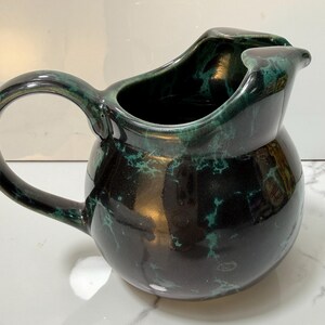 May include: A ceramic pitcher with a dark green and black marbled glaze. The pitcher has a rounded body, a curved handle, and a spout. The interior is black. This decorative pitcher is suitable for serving beverages.