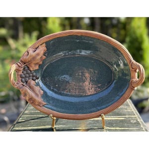 May include: Oval ceramic serving dish with a dark teal interior and a brown, textured rim. The dish features a detailed relief of grape leaves and grapes on one side and handles on each end. The dish is displayed on a gold stand.