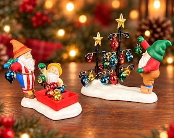 May include: Two ceramic figurines on white snowy bases. One features two bearded figures in colorful hats and outfits with a red crate of small metallic ornaments. The other shows a bearded figure in a green hat decorating a black metal tree with star toppers and hanging metallic ornaments.