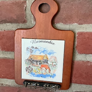 Peut inclure: Un porte-clés mural en bois avec un carreau en céramique carré. Le carreau présente une illustration colorée d'une maison et d'un cheval avec le texte Normandie. Une barre en métal noir avec quatre crochets est fixée sur le bord inférieur.