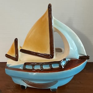 Op de afbeelding: Een keramische zeilbootplanter met een lichtblauwe romp, bruin dek en een beige zeil. Het zeil heeft een donkerbruine rand. De planter heeft een uitsparing in het midden en is ontworpen voor kleine planten of bloemen. Ongeveer 20 cm lang.
