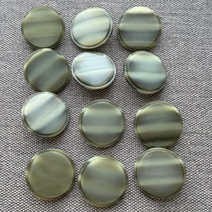 May include: Twelve circular buttons with a pearlescent, striped design in shades of grey, white, and gold. The buttons have a metallic sheen. They are arranged on a textured, grey surface. Each button is approximately 2.5 cm in diameter.